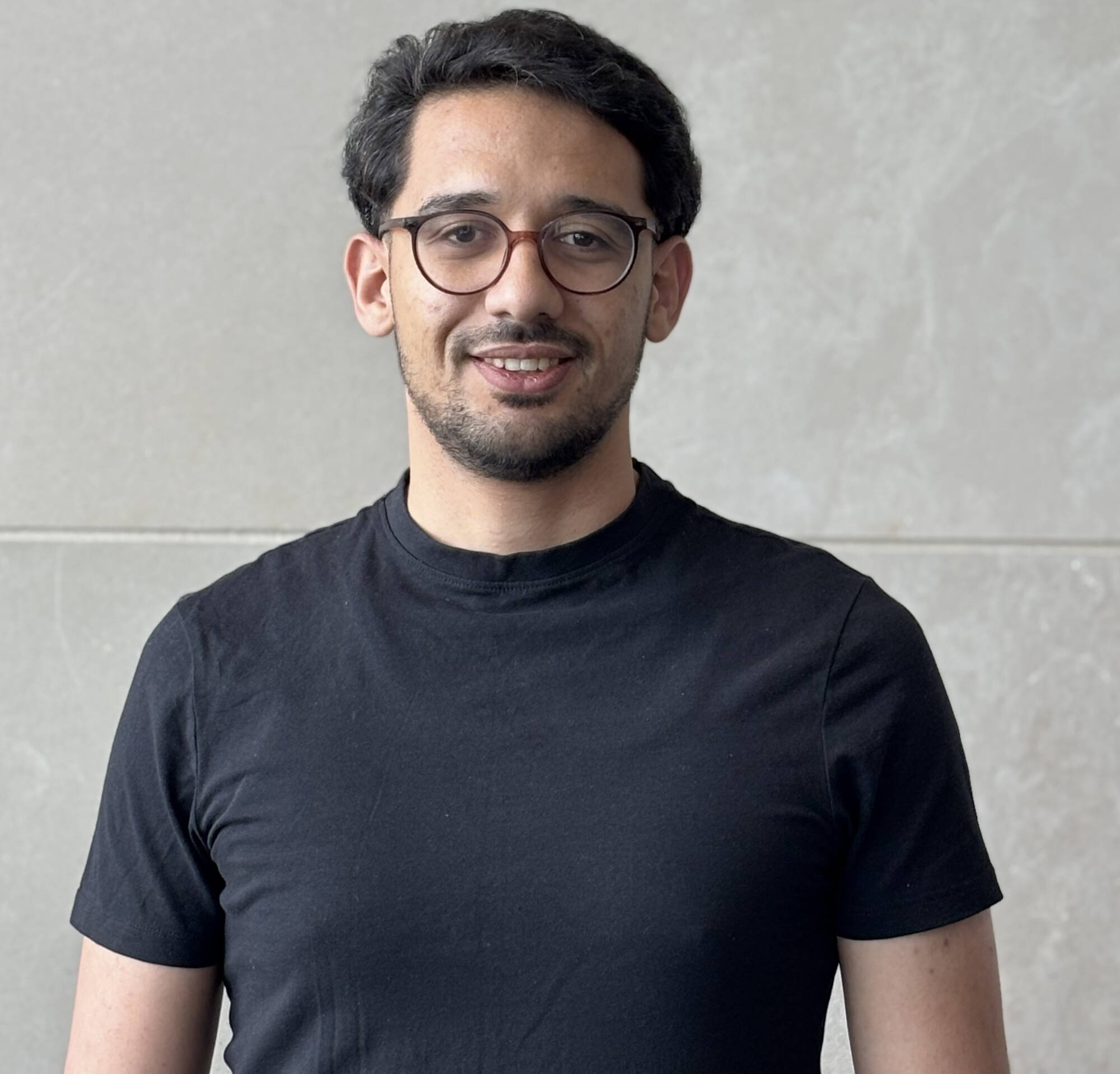 Malek Garrach, is a wearing a black  short sleeved Tshirt with a pair of spectacles on. Behind him is a gray wall